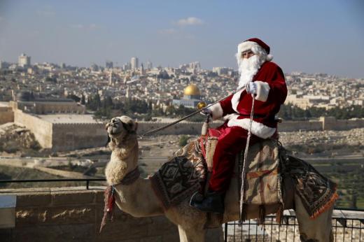 Israeli-Arab Issa Kassissieh wears a Santa Claus costume as he rides a camel and poses for the media during an annual Christmas tree distribution by the Jerusalem municipality on the Mount of Olives in Jerusalem, December 21, 2015. The Dome of the Rock is seen in the background. REUTERS/Ammar Awad